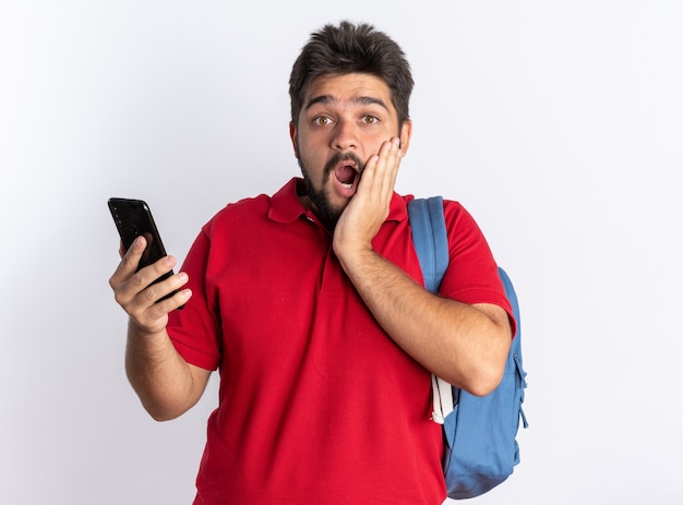 Chico joven estudiante barbudo en polo rojo con mochila sosteniendo smartphone mirando asombrado y sorprendido de pie