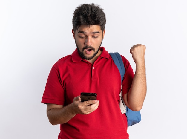 Chico joven estudiante barbudo en polo rojo con mochila sosteniendo smartphone mirándolo feliz y sorprendido de pie