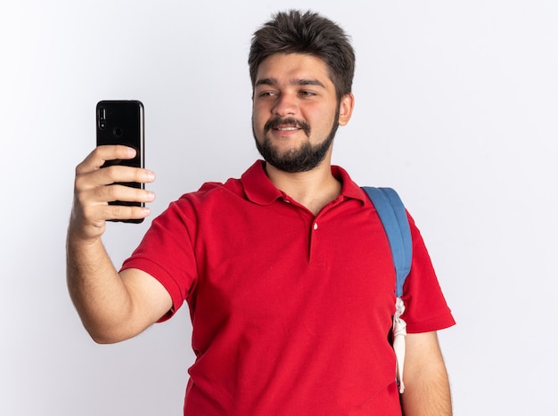 Chico joven estudiante barbudo en polo rojo con mochila sosteniendo smartphone mirándolo sonriendo alegremente de pie sobre la pared blanca