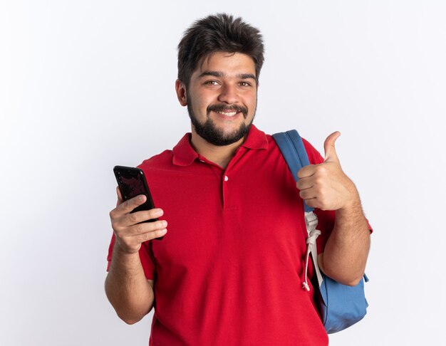 Chico joven estudiante barbudo en polo rojo con mochila sosteniendo smartphone sonriendo alegremente mostrando los pulgares para arriba de pie sobre la pared blanca