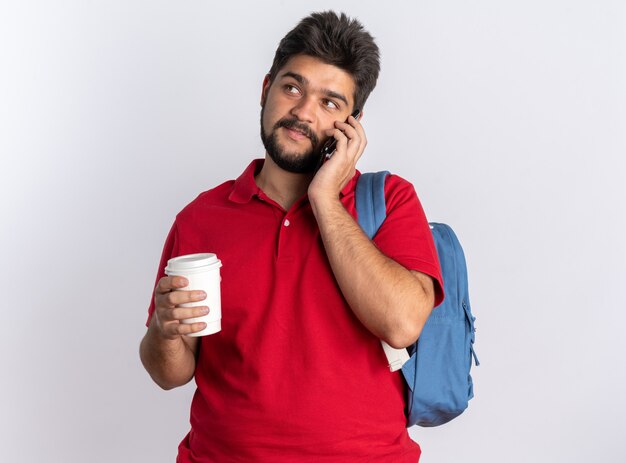 Chico joven estudiante barbudo en polo rojo con mochila sosteniendo la taza de café sonriendo confiado mientras habla por teléfono móvil de pie sobre fondo blanco.