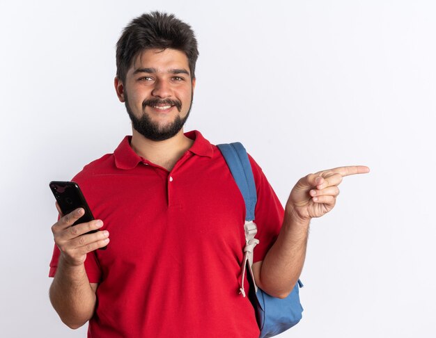 Chico joven estudiante barbudo en polo rojo con mochila sosteniendo teléfono inteligente apuntando con el dedo índice hacia el lado sonriendo confiado de pie sobre la pared blanca