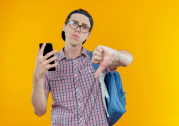 Chico joven estudiante disgustado con bolsa trasera y gafas y gorra sosteniendo el teléfono con el pulgar hacia abajo en blanco