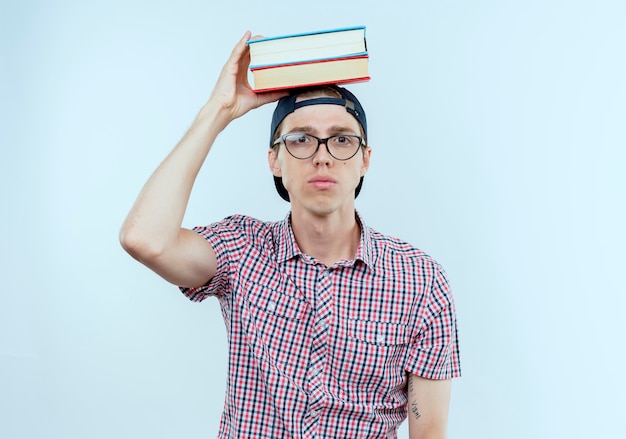 Chico joven estudiante con gafas y gorra poniendo libros en la cabeza.