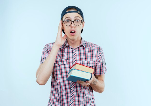 Chico joven estudiante sorprendido con bolsa trasera y gafas y gorra sosteniendo libros en blanco