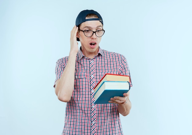 Chico joven estudiante sorprendido con bolsa trasera y gafas y gorra sosteniendo y mirando libros en blanco