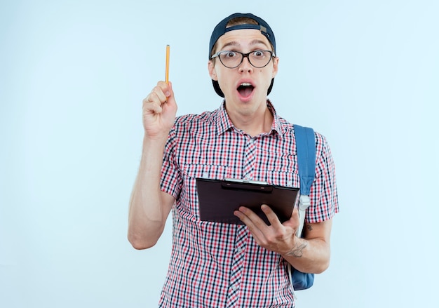 Chico joven estudiante sorprendido con bolsa trasera y gafas y gorra sosteniendo el portapapeles y levantando la pluma en blanco