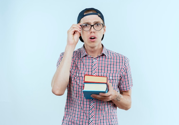 Chico joven estudiante sorprendido con bolso trasero y gafas y gorra sosteniendo libros y poniendo la mano en vasos aislados en la pared blanca