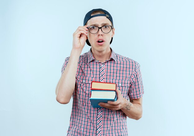 Chico joven estudiante sorprendido con bolso trasero y gafas y gorra sosteniendo libros y poniendo la mano en vasos aislados en la pared blanca