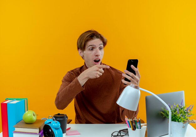 Chico joven estudiante sorprendido sentado en el escritorio con herramientas escolares sosteniendo y apunta al teléfono en amarillo
