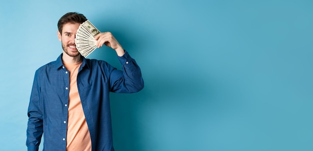 Chico joven feliz cubriendo la mitad de la cara con dólares y sonriendo ganando el dinero del premio de pie en azul