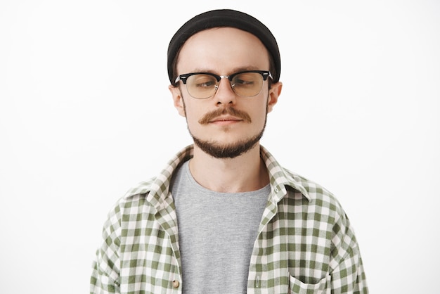 Chico joven guapo e infantil juguetón con bigote y barba con gafas y gorro negro de moda mirando la punta de la nariz, entrecerrar los ojos y hacer una cara divertida mientras tontea sin tener nada que hacer estando aburrido