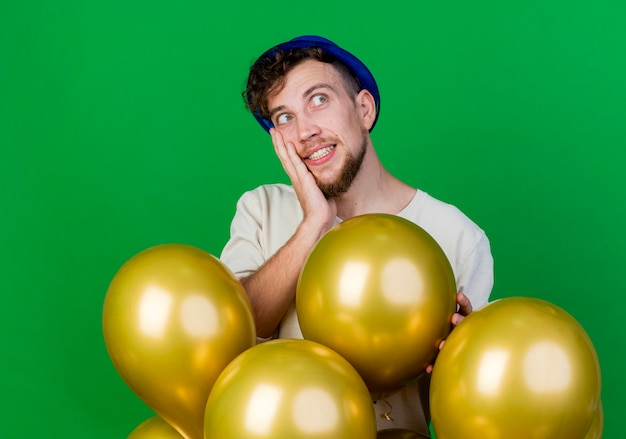 Chico joven guapo partido eslavo con sombrero de fiesta de pie detrás de globos poniendo la mano en la cara mirando de lado sumergiéndose en sueños aislados sobre fondo verde