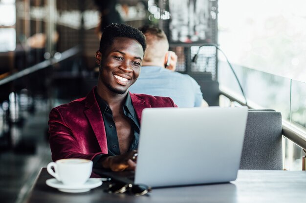 Un chico joven y guapo de piel oscura con un traje sentado en un café y una computadora portátil.