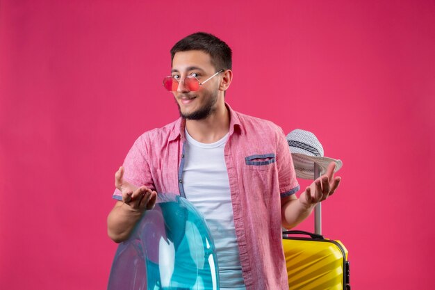 Chico joven guapo viajero con gafas de sol con anillo inflable de pie con maleta de viaje mirando a cámara sonriendo astutamente con los brazos levantados sobre fondo rosa