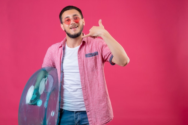 Chico joven guapo viajero con gafas de sol sosteniendo anillo inflable mirando a la cámara sonriendo alegremente haciendo gesto de llamarme de pie sobre fondo rosa