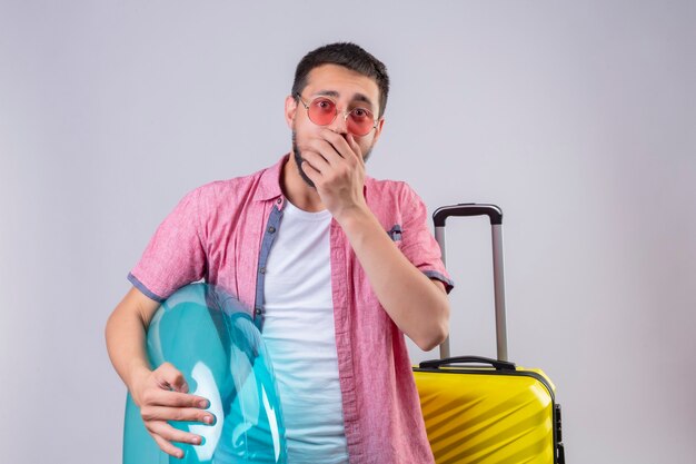 Foto gratuita chico joven guapo viajero con gafas de sol sosteniendo anillo inflable mirando a cámara sorprendido y asombrado cubriendo la boca con la mano de pie con maleta de viaje sobre fondo blanco