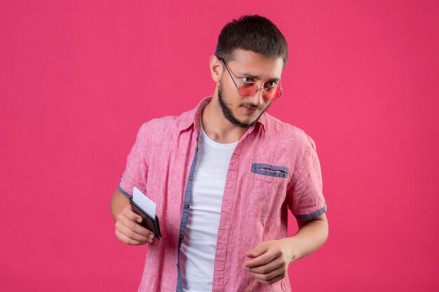 Chico joven guapo viajero con gafas de sol sosteniendo boletos mirando a un lado con expresión de confianza de pie sobre fondo rosa