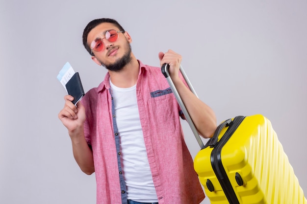 Chico joven guapo viajero con gafas de sol sosteniendo maleta y billetes de avión mirando a la cámara con sonrisa de confianza de pie sobre fondo blanco