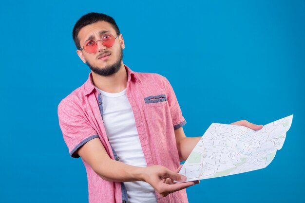 Chico joven guapo viajero con gafas de sol sosteniendo mapa mirando decepcionado de pie sobre fondo azul.