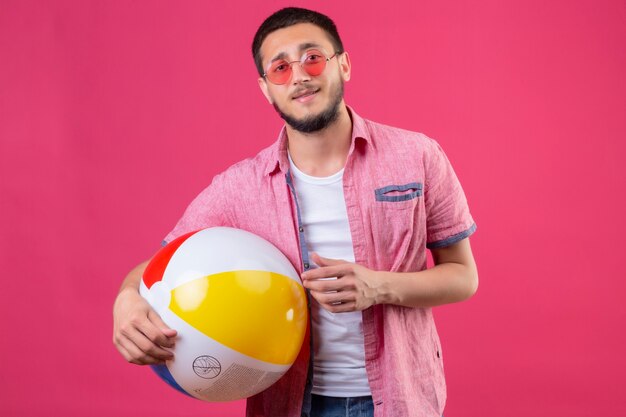 Chico joven guapo viajero con gafas de sol sosteniendo una pelota inflable mirando a la cámara con una sonrisa de confianza en la cara de pie sobre fondo rosa