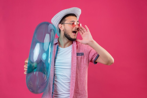 Chico joven guapo viajero con sombrero de verano con gafas de sol con anillo inflable gritando o llamando a alguien con la mano cerca de la boca de pie sobre fondo rosa