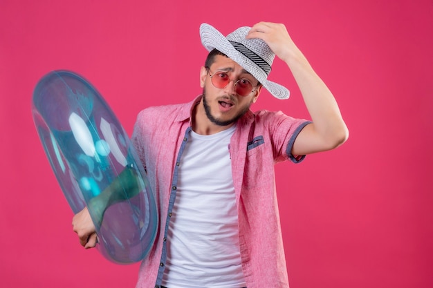 Chico joven guapo viajero con sombrero de verano con gafas de sol sosteniendo un anillo inflable despistado y confundido tocando su sombrero sin respuesta de pie sobre fondo rosa