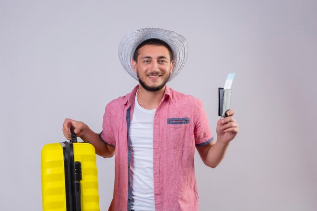 Chico joven guapo viajero con sombrero de verano con maleta y billetes de avión mirando a cámara con gran sonrisa en la cara feliz y positivo de pie sobre fondo blanco