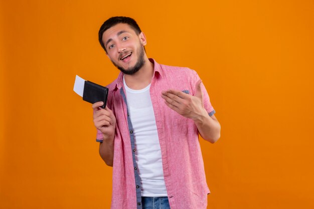 Chico joven guapo viajero sosteniendo billetes de avión apuntando con el brazo de la mano a ellos feliz y positivo mirando a la cámara sonriendo de pie sobre fondo naranja