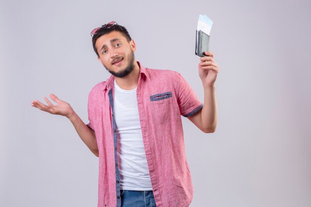 Chico joven guapo viajero sosteniendo billetes de avión despistado y confundido de pie con los brazos levantados sin respuesta sobre fondo blanco