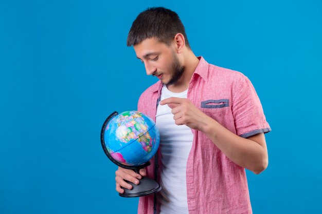 Chico joven guapo viajero sosteniendo globo mirándolo con seria expresión de confianza de pie sobre fondo azul.