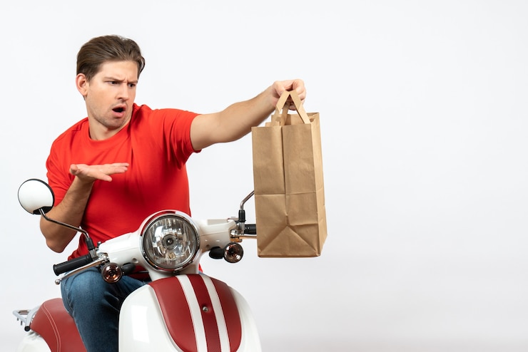 Foto gratuito chico joven mensajero emocional confundido en uniforme rojo sentado en scooter sosteniendo una bolsa de papel en la pared blanca
