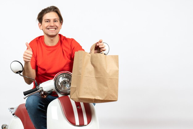 Chico joven mensajero feliz en uniforme rojo sentado en scooter dando bolsa de papel haciendo gesto ok en la pared blanca