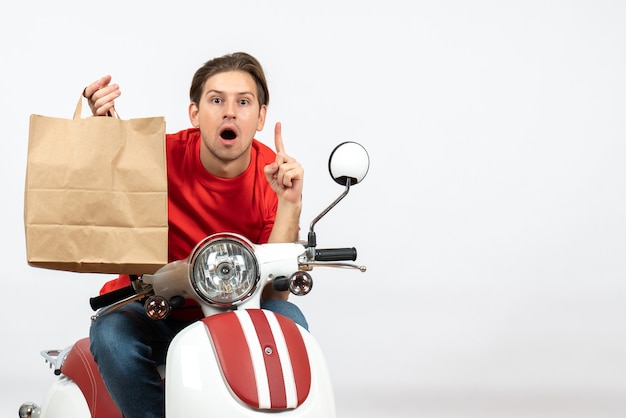 Chico joven mensajero sorprendido en uniforme rojo sentado en scooter sosteniendo una bolsa de papel apuntando hacia arriba en la pared blanca