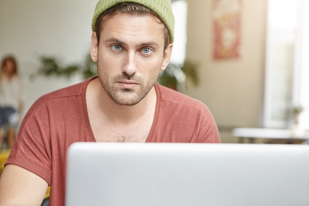 Chico joven con ojos azules y barba se ve con confianza mientras se sienta frente a la computadora portátil abierta, revisa el correo electrónico o navega por las redes sociales en línea