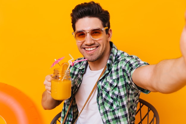 Chico joven de pelo oscuro con camisa verde y gafas naranjas disfruta de un cóctel y toma selfie en un espacio aislado.