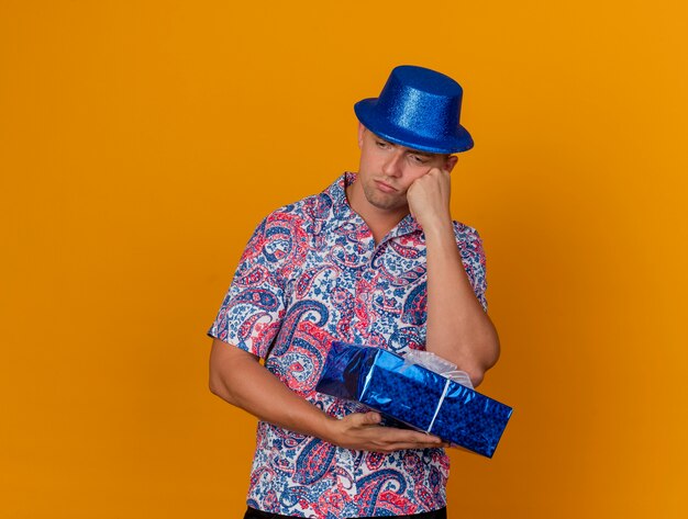 Chico joven pensativo mirando hacia abajo con sombrero azul sosteniendo una caja de regalo y poniendo la mano en la mejilla aislado en naranja