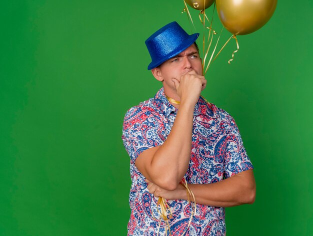 Chico joven pensativo mirando al lado con sombrero azul sosteniendo globos atados alrededor del cuello y poniendo la mano en la boca aislada en verde