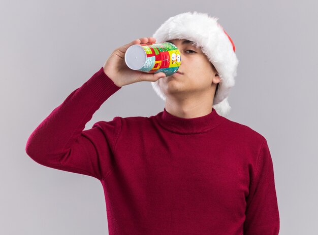 Chico joven con sombrero de Navidad sosteniendo la copa de Navidad y bebidas aislado en la pared blanca