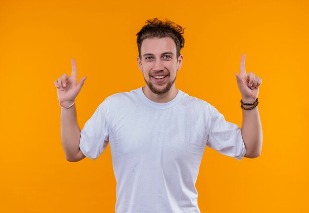 Chico joven sonriente con camiseta blanca apunta hacia arriba sobre fondo naranja aislado