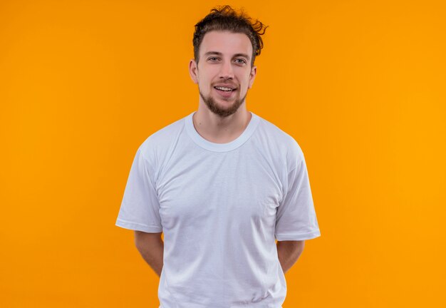 Chico joven sonriente con camiseta blanca puso su mano en la espalda sobre fondo naranja aislado