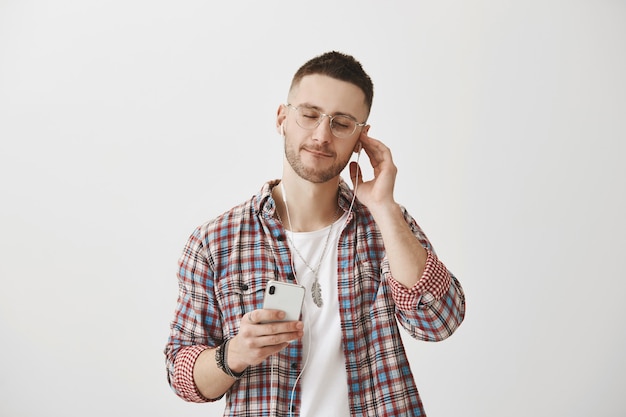 Chico joven sonriente despreocupado con gafas posando con su teléfono