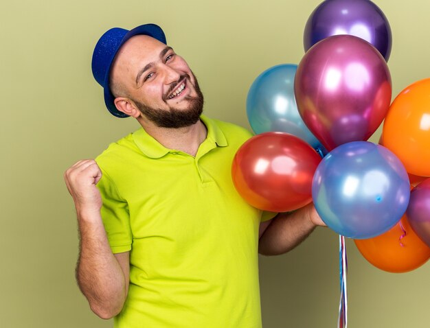 Chico joven sonriente con gorro de fiesta sosteniendo globos mostrando gesto de sí