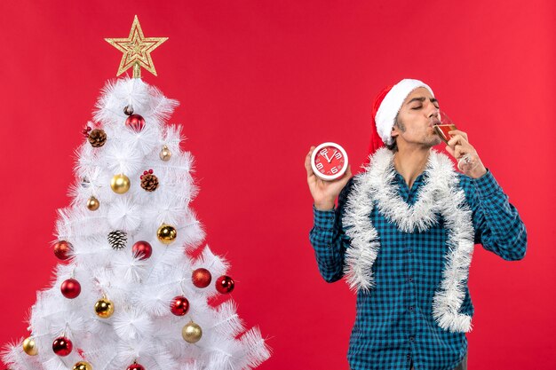 Chico joven sonriente con sombrero de santa claus y bebiendo una copa de vino y sosteniendo el reloj de pie cerca del árbol de Navidad en rojo