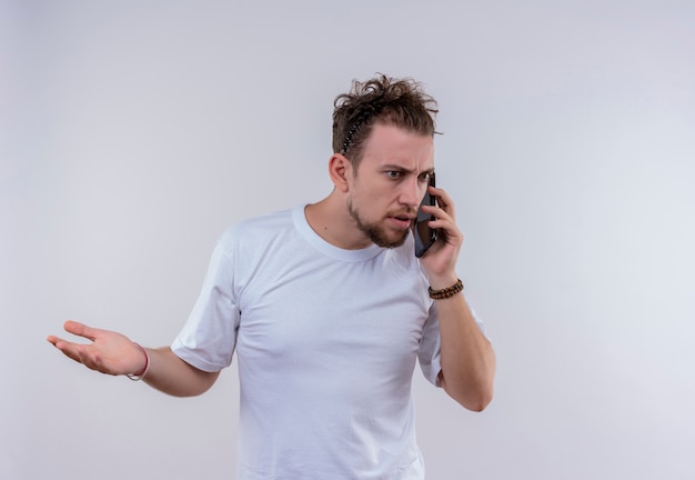 Chico joven sorprendido con camiseta blanca habla por teléfono levantando la mano sobre fondo blanco aislado