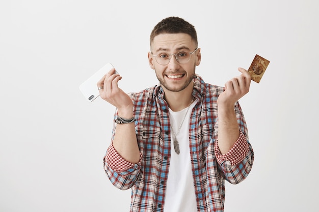 Chico joven sorprendido con gafas posando con su teléfono y tarjeta