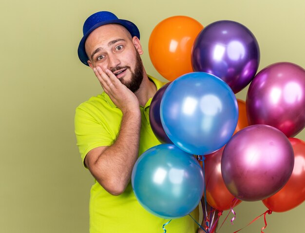 Chico joven sorprendido con gorro de fiesta sosteniendo globos poniendo la mano en la mejilla