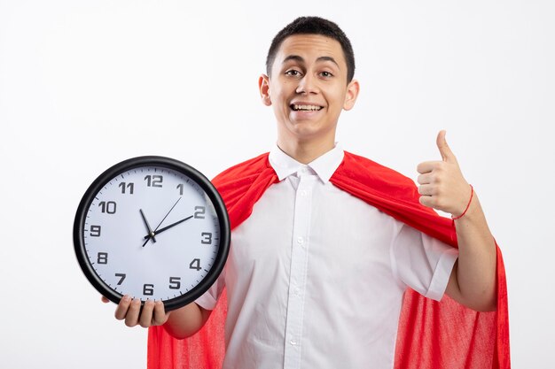 Chico joven superhéroe alegre en capa roja sosteniendo el reloj mirando a la cámara mostrando el pulgar hacia arriba aislado sobre fondo blanco.