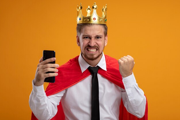 Chico joven superhéroe alegre con corbata y corona sosteniendo el teléfono y mostrando un gesto de sí aislado sobre fondo naranja