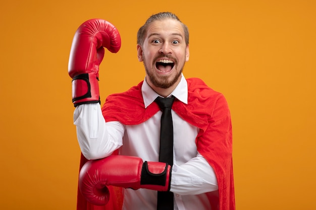 Chico joven superhéroe alegre con corbata y guantes de boxeo levantando el puño aislado en naranja